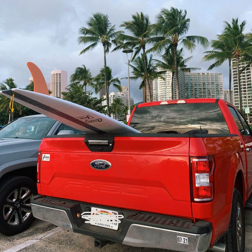 Red rental car in the parking lot of Waikiki Beach with a surfboard in the trunk and palm trees in the background