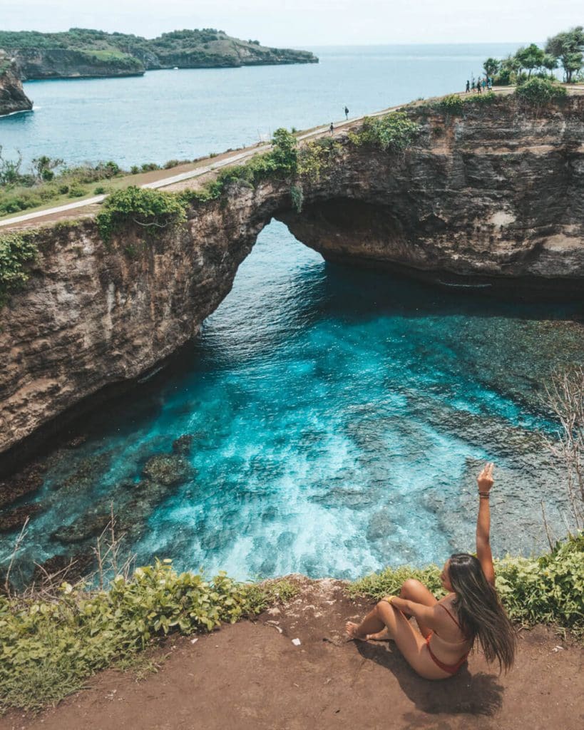 Girl sitting in front of Broken Beach at Nusa Penida with clear blue waters and a natural stone bridge