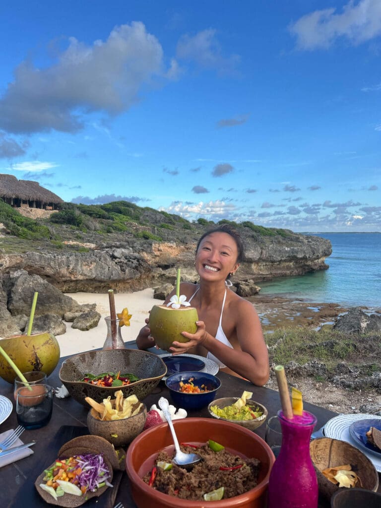 Danielle from The Wanderlover smiling at the camera holding a coconut with a frangipani flower and a spread of delicious food in front of her. There are cliffs and nature inspired huts behind here above a clear blue lagoon.