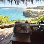 Laptop open to The Wanderlover Website on a wooden balcony overlooking bright blue waters and green fauna in Rote, Indonesia.